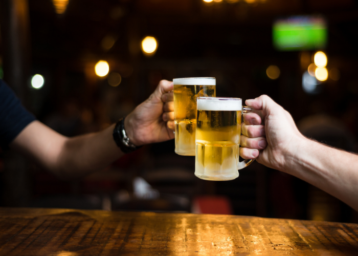 fotografia mostrando duas mãos segurando canecas de chopp e fazendo um brinde sob uma mesa de madeira