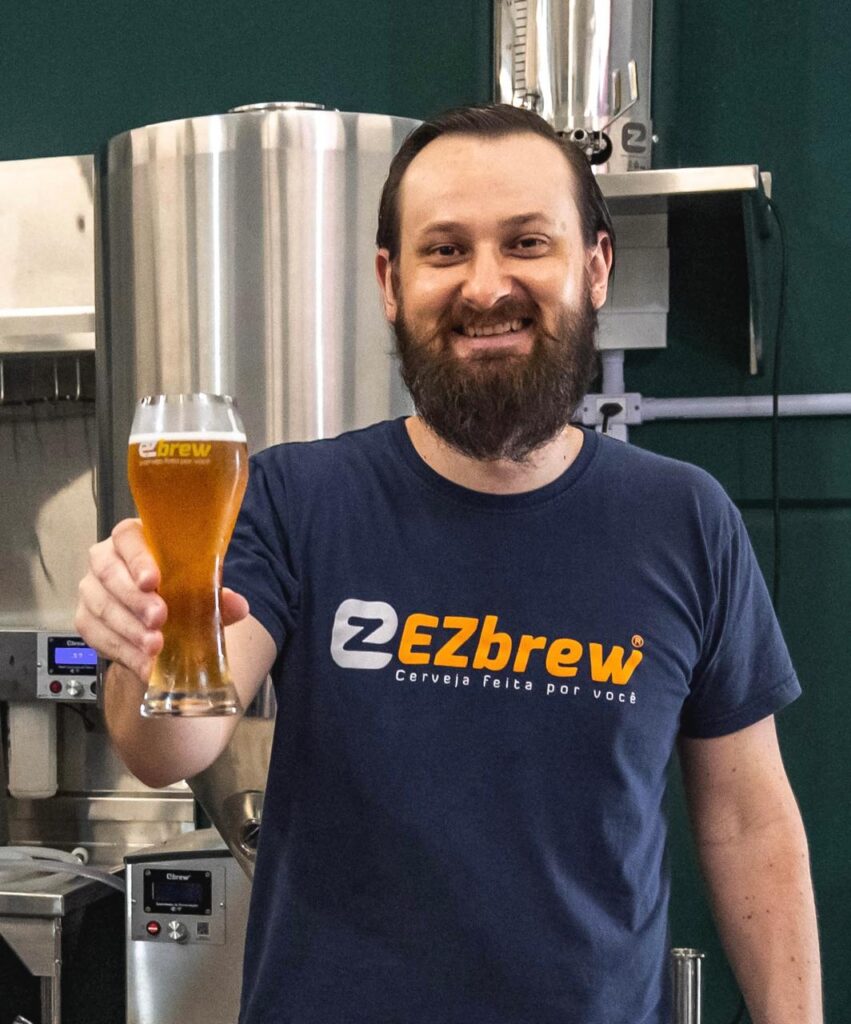 Homem sorridente segurando um copo de cerveja artesanal clara, vestindo uma camiseta da EZbrew, em uma cervejaria com tanques de fundo.